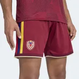 Short de fútbol Adidas, versión Fan, color granate (similar al rojo oscuro), con detalles en amarillo en los bordes. Presenta el escudo de la Federación Venezolana de Fútbol (FVF) bordado en el muslo izquierdo.