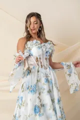 Vestido largo de fiesta en gasa de seda natural color blanco con estampado de flores celestes y verdes. Corsé strapless con ballenas y detalle de flores en escote y mangas caídas. Espalda cruzada y falda amplia con tajo delantero.