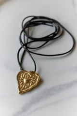 Collar con dije de corazón en acero quirúrgico, con textura irregular y relieve, montado sobre un tiento encerado negro.