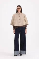 Piloto corto estilo camisa en color beige, con cuello clásico, cierre frontal de botones y mangas anchas con detalle de trabas en los puños.