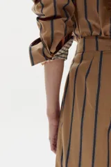 Camisa boxy de popelina de algodón color tostado con diseño de rayas verticales oscuras, cuello camisero, botones frontales y mangas largas con puños estampados a cuadros.