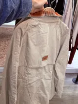 Gabardina de corte clásico en color verde militar, con solapas amplias y puños con detalles en estampado de cuadros escoceses en tonos azul, blanco y negro. Presenta cierre frontal cruzado con botones y bolsillos laterales.