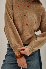 Sweater de punto color beige melange con bordado de pequeños corazones rojos distribuidos por toda la prenda. Posee cuello redondo, mangas largas y terminaciones acanaladas en puños y ruedo.