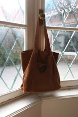 Cartera tipo tote de gamuza color marrón, con diseño amplio y relajado. Presenta un bolsillo frontal externo con cierre de broche metálico y una hebilla decorativa circular, además de un detalle de borla colgante en el lateral.