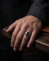 Anillo de acero tipo sello con piedra central redonda color negro y detalles de guarda griega en los laterales.