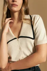 Top de tejido color beige claro, manga corta, escote en V con cierre frontal de botones y ribetes negros en el cuello y el borde del escote.