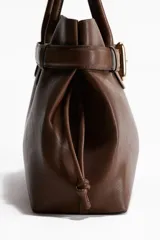 Bolso tote marrón con doble asa corta, cierre superior con cordón ajustable y cinturón decorativo con hebilla metálica dorada.