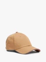 Gorra de béisbol color beige con detalle distintivo de franja tricolor (rojo, blanco y azul) en el borde de la visera. Tiene logo metálico de Tommy Hilfiger en el frente y hebilla metálica ajustable en la parte trasera.