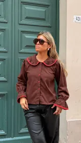 Camisa de color marrón chocolate con cuello Peter Pan y puños con volados, ambos detalles resaltados con un ribete en color rojo. Presenta cierre frontal con botones rojos.