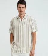 Camisa de hombre confeccionada en 100% algodón con textura, de manga corta y cuello inglés. Presenta un diseño de rayas verticales en tonos beige, blanco y marrón, con cierre frontal de botones.