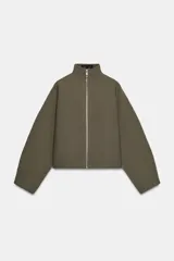 Chaqueta corta de tejido grueso color verde oliva, con cuello alto y cierre frontal completo de cremallera plateada. Presenta un corte amplio y mangas largas caídas.