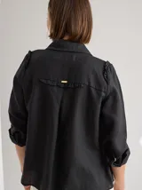 Camisa de lino color negro, con cuello clásico, cierre frontal de botones de madera y detalle de volados en los hombros. Presenta costuras a la vista en tono contrastante.