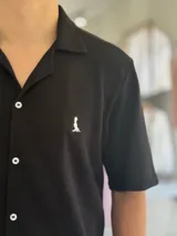 Camisa de manga corta color blanco marfil, confeccionada en tejido texturizado con cuello camisero, cierre frontal de botones y un pequeño bordado oscuro en el pecho.