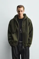 Sudadera de corte relajado confeccionada en tejido técnico con elasticidad y efecto neopreno. Presenta capucha ajustable con cordones, cierre frontal de cremallera de doble dirección, bolsillos tipo canguro y terminaciones en rib.