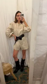 Mono corto de lino color beige, con cuello camisero, cierre frontal, mangas largas abullonadas y cinto ajustable en la cintura.