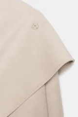 Abrigo de mezcla de lana color beige claro, de corte oversize y largo medio. Presenta un cuello solapa envolvente con detalle de pañuelo integrado y manga larga. Cierre frontal cruzado con botones ocultos.