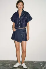 Camisa corta de denim de manga corta, con cuello solapa, escote en V y bolsillos delanteros de plastrón. Cuenta con costuras en contraste y cierre frontal con botones metálicos.