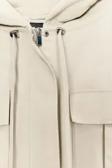 Cazadora color beige con capucha ajustable, manga larga con puño elástico, bolsillos frontales con solapa, bajo ajustable y cierre frontal con cremallera y botones a presión.