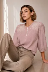 Blusa a rayas verticales en tonos rosados y blancos, con cuello redondo y cierre frontal parcial con botones. Presenta delicados detalles bordados en los hombros y mangas tres cuartos abullonadas con puños elásticos.