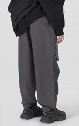 Pantalón cargo oversize de color gris, confeccionado en tejido técnico ripstop. Cuenta con cintura elástica con presillas, cierre frontal con broche técnico, múltiples bolsillos amplios y sistema de ajuste con broches snap en los tobillos.