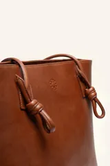 Cartera tote de cuero suave color marrón, con diseño amplio y líneas simples, con tiras de hombro decoradas con nudos artesanales y pequeño logo grabado en el centro.