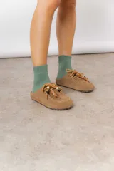 Zueco de cuero gamuzado color tostado, con cordones decorativos adornados con cuentas de madera y piezas envueltas en cuero. Diseño tipo mocasín con costuras visibles en la parte superior y abierto en la parte trasera.