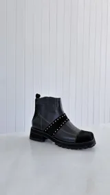 Botín de cuero negro con diseño de paneles combinados en cuero liso y gamuza. Presenta una banda diagonal con tachas metálicas plateadas, costuras decorativas en zigzag y suela track robusta.