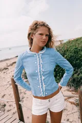 Chaqueta de popelín a rayas celestes y blancas, con cuello redondo y mangas largas. Presenta detalles de encaje a contraste en el cuello, los puños y la botonadura frontal, la cual cuenta con cierre de botones.