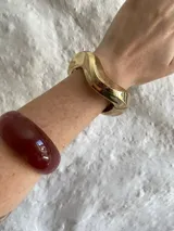 Brazalete rígido de diseño orgánico y asimétrico con acabado metálico brillante en tono dorado. Cuenta con un sistema de cierre de bisagra integrado.