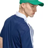 Remera deportiva Adidas Originals azul marino de manga corta, con cuello polo y solapa en V de color blanco. Presenta las icónicas tres tiras blancas en los hombros, el logotipo clásico de la marca en el pecho y un sutil patrón de rayas verticales tejidas.