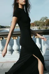 Vestido largo negro de cuello redondo y manga corta, con detalle de drapeado lateral, bajo con abertura lateral y cierre de cremallera en la espalda.