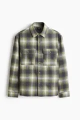 Sobrecamisa de franela suave con cuello inglés, bolsillos superiores con solapa y cierre frontal de botones. Presenta un diseño de cuadros en tonos verdes, grises y blancos, con mangas largas y bajo recto.