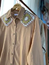 Camisa tostada de manga larga con cuello clásico decorado con apliques de crochet en tonos marrones, blancos y verdes, cierre frontal de botones y diseño holgado.