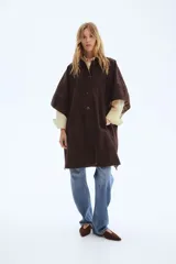 Abrigo oversized de sarga color marrón con cuello de cuero sintético. Modelo con botones en el frente, bolsillos laterales discretos, mangas tipo capa y aberturas laterales con botón.