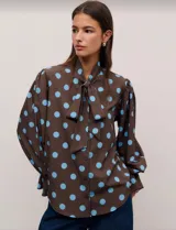 Camisa de manga larga con estampado de lunares celestes sobre fondo marrón. Presenta cuello con lazo para anudar, mangas amplias con puños acampanados y cierre frontal con botones.