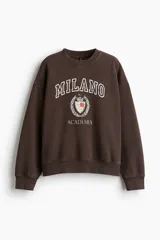 Buzo de tejido de punto color marrón oscuro con efecto lavado, cuello redondo y hombros caídos. Presenta un estampado frontal con el texto "MILANO" en tipografía universitaria, un escudo con la inscripción "193" y la palabra "ACADEMIA" debajo.