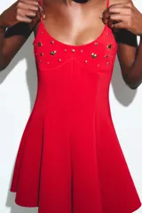 Vestido corto rojo, ajustado en el busto y con falda acampanada. Tiene tirantes finos ajustables y apliques metálicos dorados en forma de corazón y redondos en el escote.