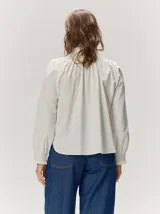 Camisa de algodón color crudo, con cuello alto fruncido, cierre frontal abotonado y mangas largas con puños elásticos fruncidos.