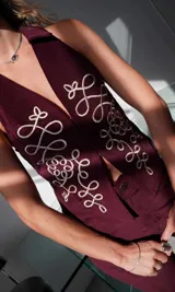Chaleco de pana color bordó, ajustado al cuerpo, con pinzas y elásticos en la espalda baja. Presenta un largo irregular, cuello clásico camisero y cierre frontal con ganchos peleteros. Tiene bordados decorativos en color crema en el frente.