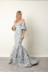 Vestido largo de fiesta color gris perla, corte sirena, con escote off-shoulder y mangas cortas con volados. Confeccionado en tul bordado con hilo al tono y leve brillo.