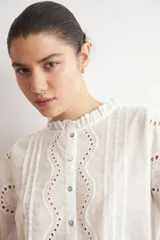 Camisa blanca de manga larga con cuello alto y volados, confeccionada en tela con bordado inglés y detalles calados verticales. Presenta puños con volados y cierre frontal con botones.