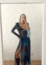 Vestido largo de microtul transparente, color azul oscuro con estampado floral en tonos azules y negros. Presenta escote pronunciado en V con volados en el borde y lazos ajustables en el centro, mangas largas con puños acampanados y volados, y abertura frontal.