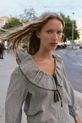 Blusa de manga larga con estampado de cuadros vichy en blanco y negro. Presenta un escote en V adornado con un volante amplio y un lazo decorativo en el centro. Las mangas terminan en puños elásticos.