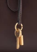 Bolso de mano de poliuretano color marrón oscuro, con doble asa para el hombro, herrajes metálicos dorados y detalle de llavero decorativo con candado y llaves.