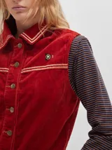 Chaleco de pana negro con cierre frontal de botones metálicos dorados. Presenta detalles de costuras en contraste en el cuello, cartera y canesú, e incluye un pequeño parche con forma de bandera en el pecho.