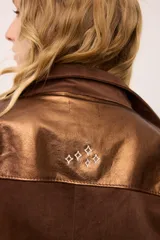 Sobrecamisa de gamuza color marrón con apliques de cuero metalizado en tono cobre en los hombros y bolsillo frontal. Presenta detalles bordados en las mangas, el bolsillo y la espalda, con un diseño de corte recto y cuello camisero.