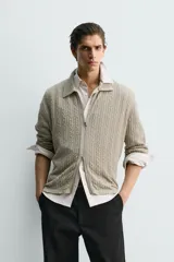 Chaqueta de punto con textura de trenzas verticales, color beige, cuello solapa y cierre frontal con cremallera.