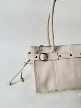 Cartera de cuero de tamaño medio con estructura rígida. Presenta una tira decorativa con tachas y hebillas metálicas en los extremos, además de lazos laterales. Cuenta con doble asa para llevar al hombro y cierre superior.