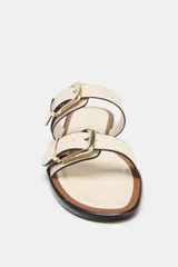 Sandalia plana de color beige con dos tiras ajustables mediante hebillas metálicas doradas. Presenta un diseño de punta redonda y suela plana con borde en contraste.