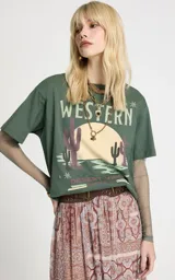 Remera de algodón color verde con estampado frontal estilo western, cuello redondo y mangas cortas. Incorpora mangas largas ajustadas de tul integradas debajo de las mangas cortas.